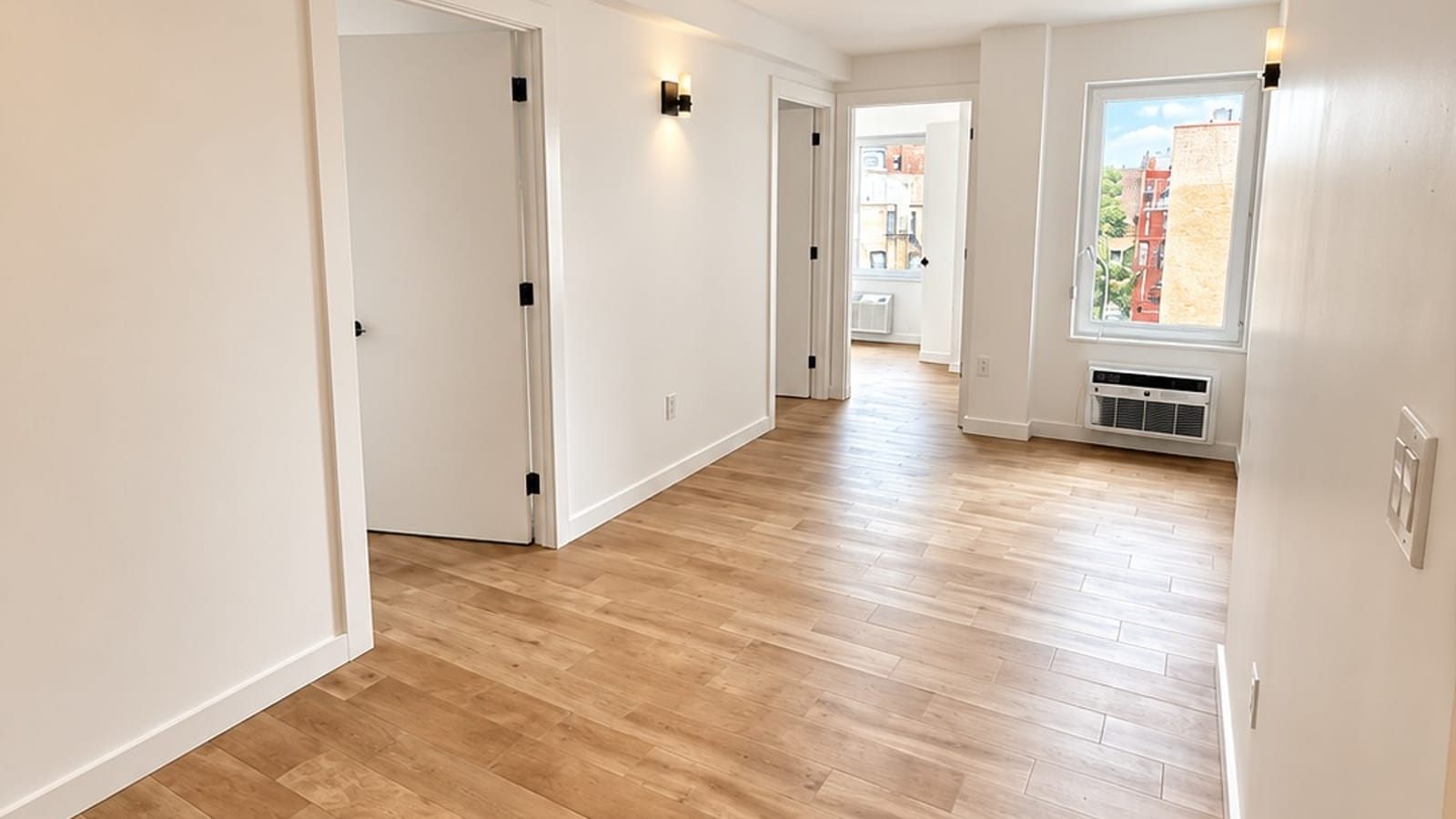 Empty apartment with wood-look floors, white walls, doorways to other rooms and a window with a wall heating and cooling unit below it