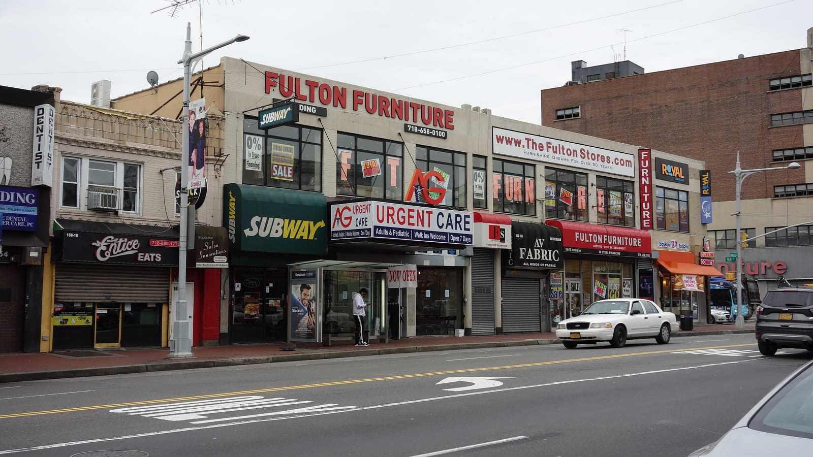 A two-story commercial building on a wide avenue with signs for a furniture store, a sandwich shop, an urgent care clinic and a fabric store, a street lamp, a bus shelter and a white car passing