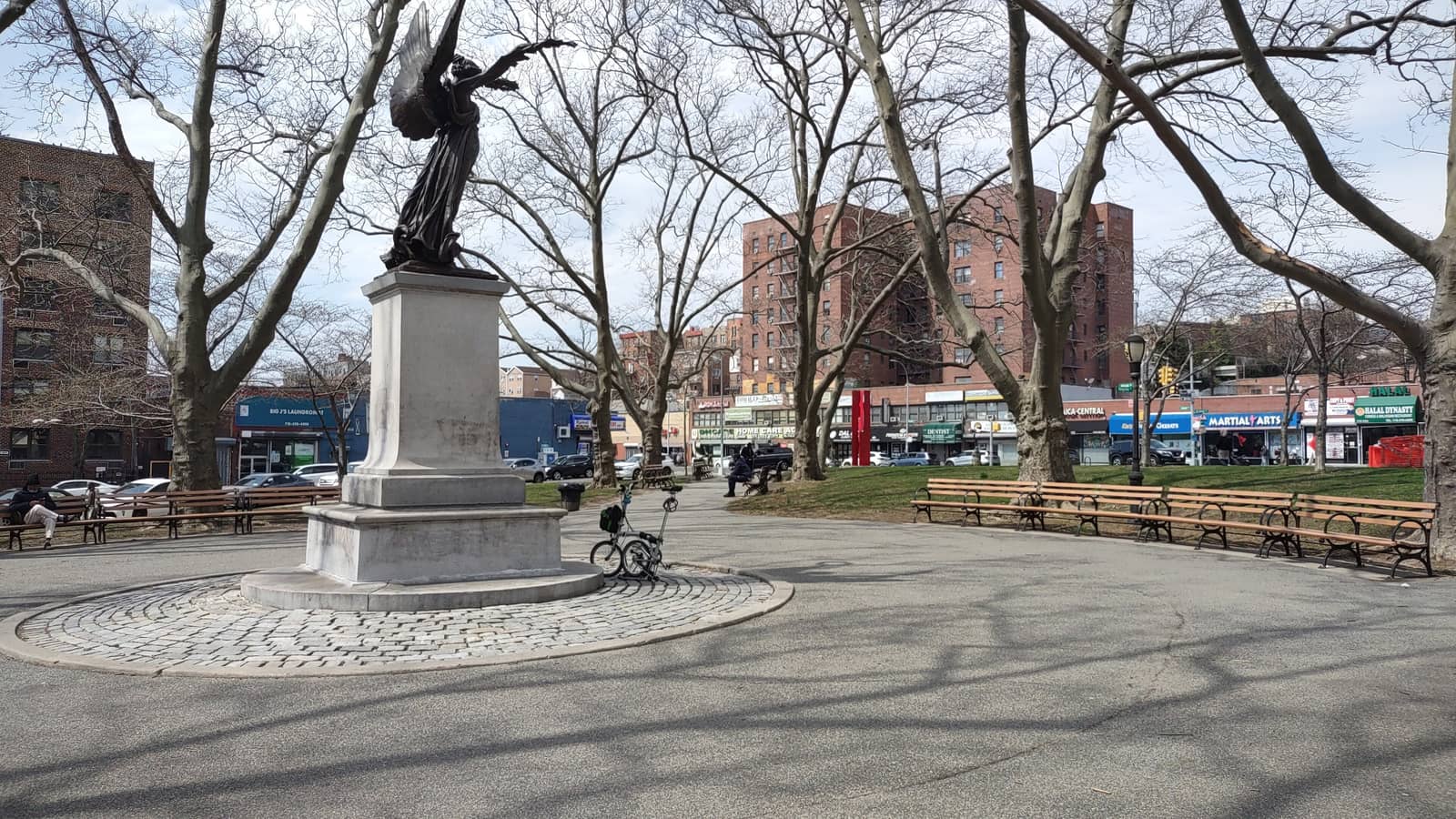 A bronze winged angel statue on a stone pedestal in a paved circle, with bare trees, long park benches, a folding bicycle and brick apartment buildings and shops behind