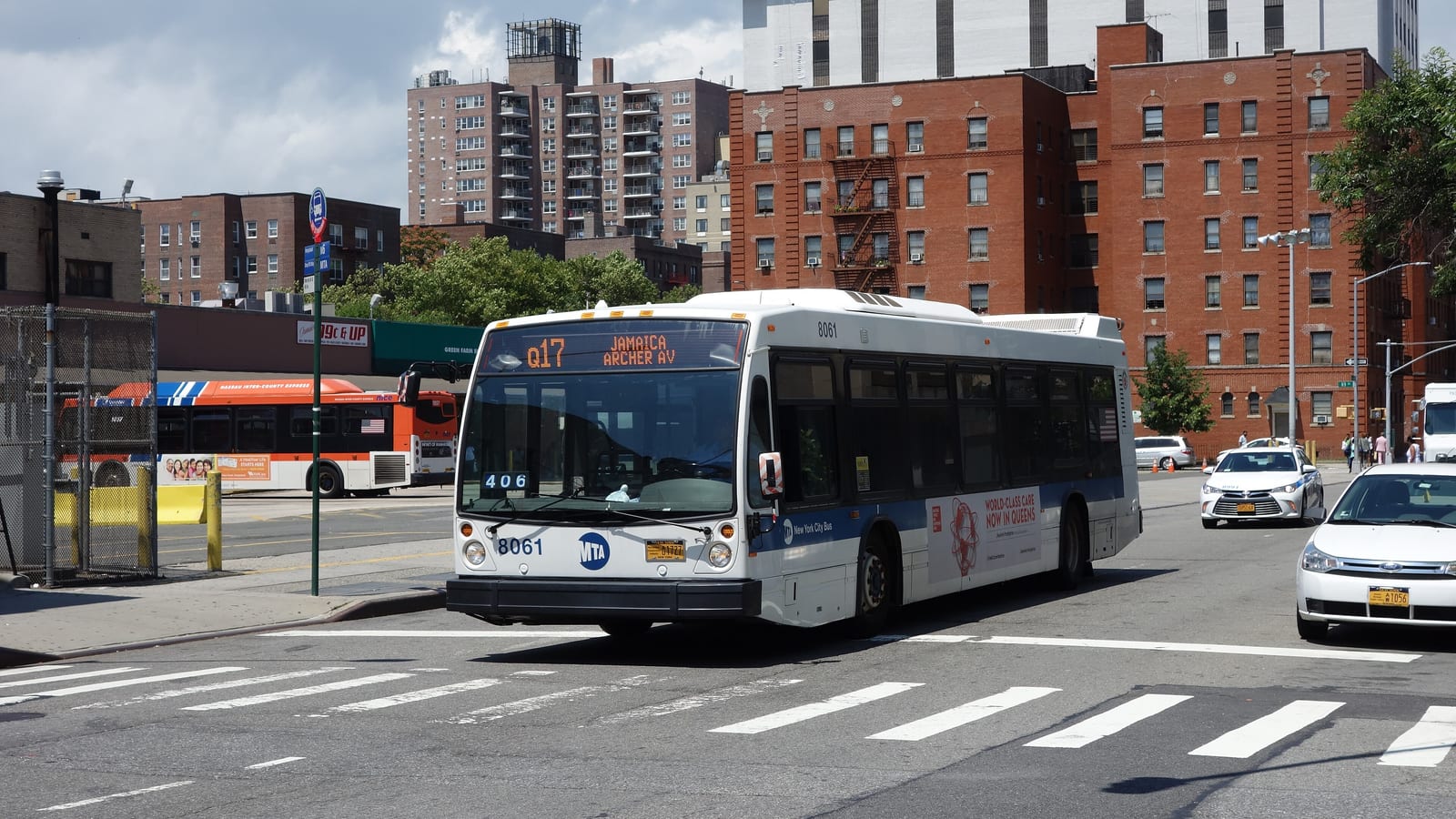 A white and blue city bus showing Q17 Jamaica Archer Av on its sign turning at a crosswalk, with an orange bus at a terminal behind, and red brick apartment buildings and a taller apartment tower in the background