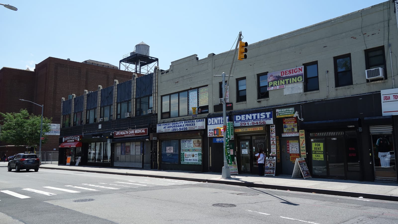 A row of two-story shop buildings on a wide street corner, with signs for dentists, a medical office and a printing shop, a traffic light on a pole, a crosswalk and a car, and a large brick building with a water tower behind
