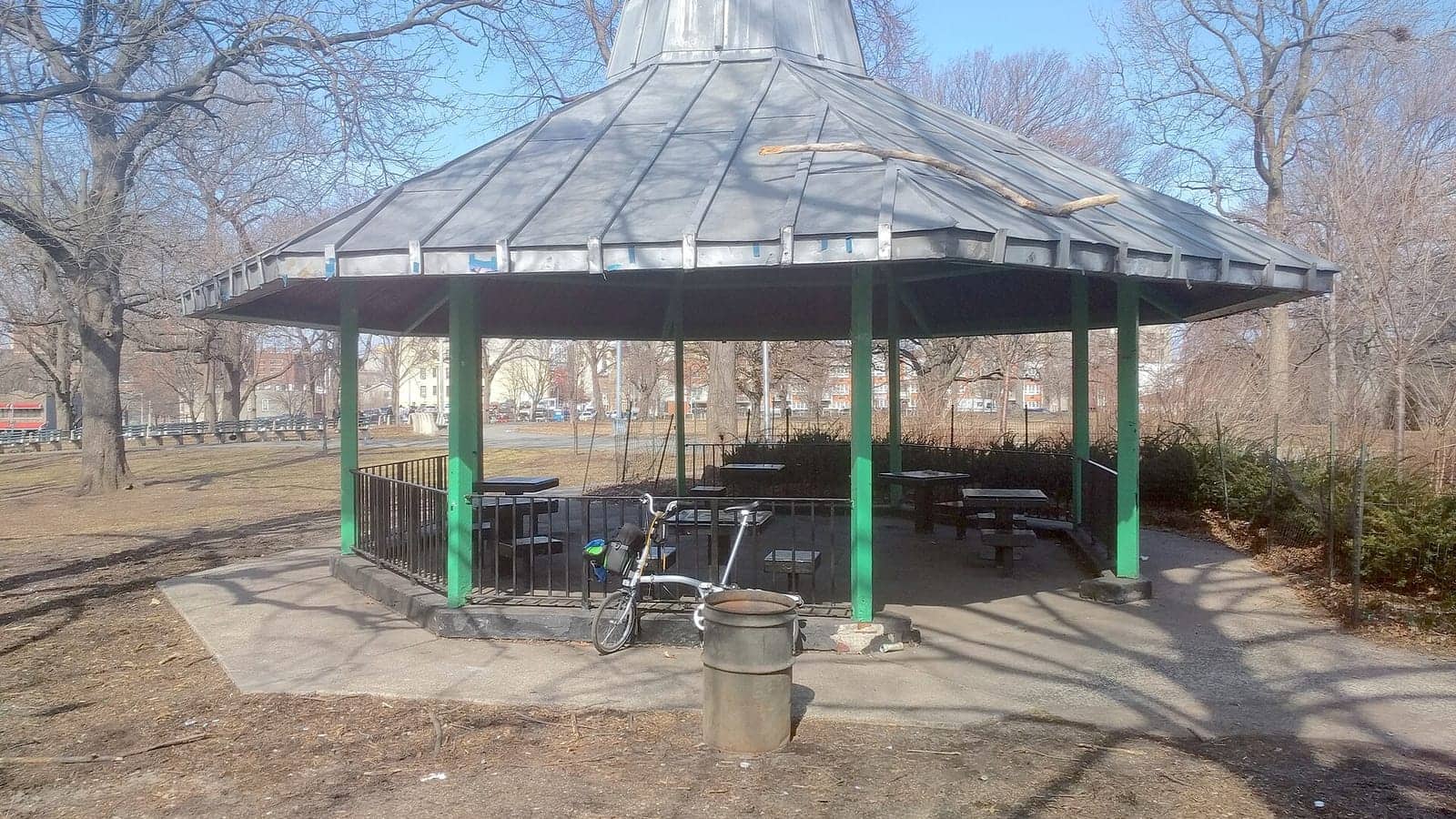 Photograph of an octagonal park pavilion with a grey metal roof on green posts, a railing around chess tables, a folding bicycle and a metal trash can in front, and bare trees and apartment buildings behind