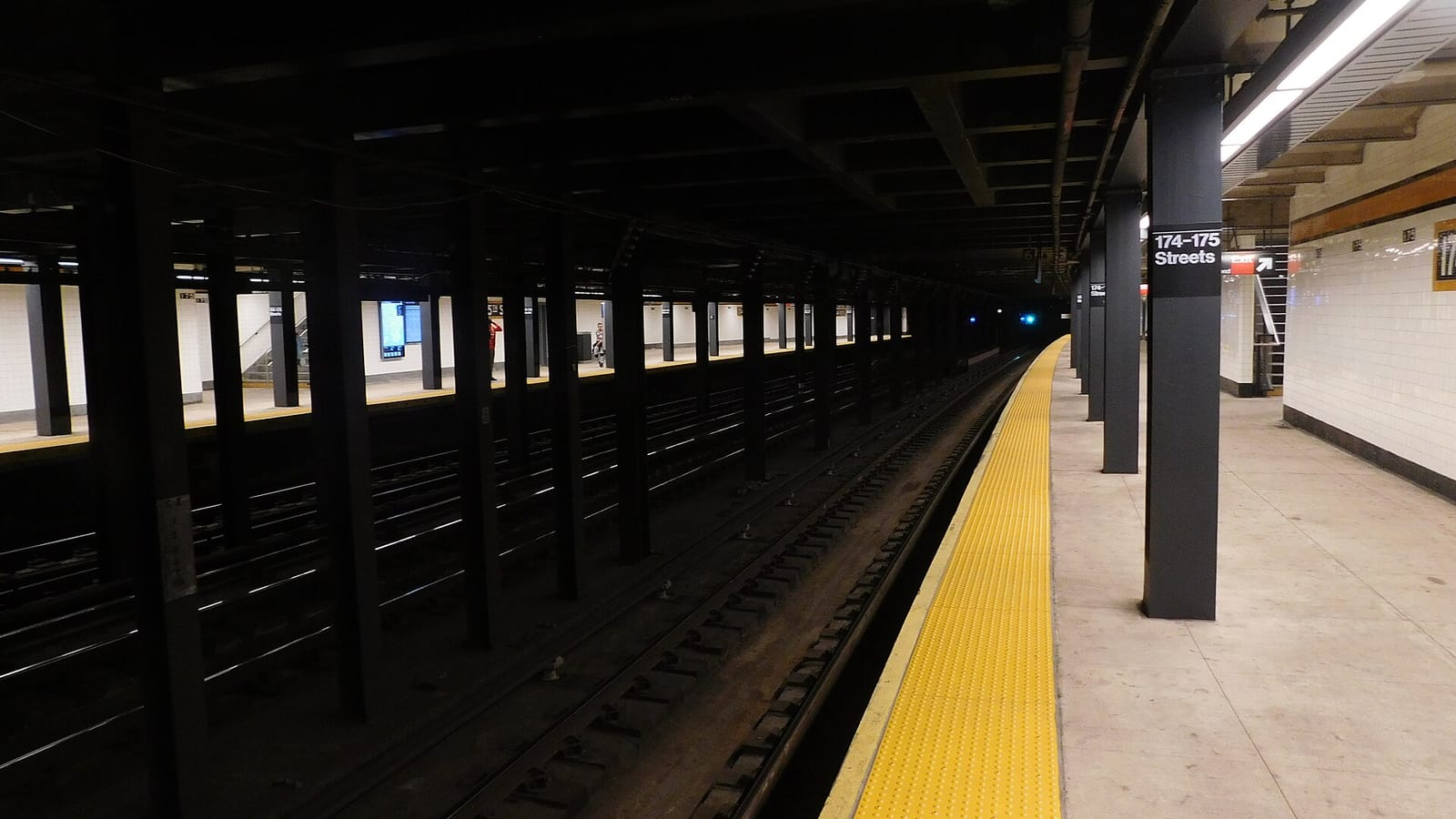 Photograph of an underground subway platform with black steel columns, one marked 174-175 Streets, a yellow tactile strip along the platform edge, tracks to the left and an opposite platform in the distance