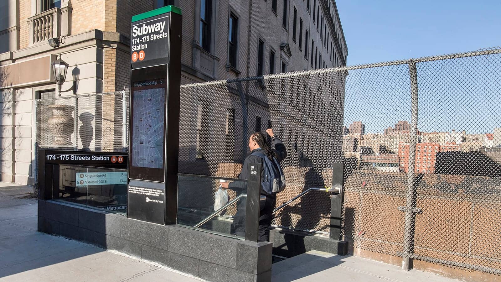 Photograph of a subway stair entrance with a tall black sign reading Subway 174-175 Streets Station with B and D route symbols and a neighborhood map, a rider walking down the stairs seen from behind, a chain-link fence and a stone building