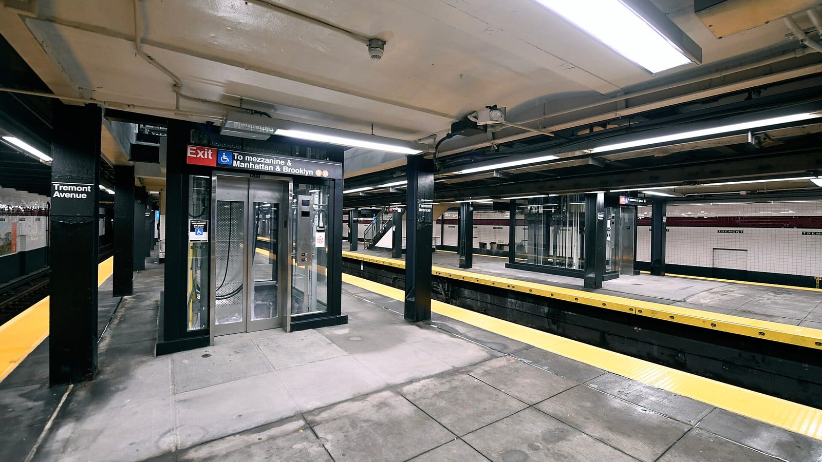 Photograph of a glass and steel elevator on an underground subway platform under a red Exit sign with a wheelchair symbol reading To mezzanine and Manhattan and Brooklyn, beside a column marked Tremont Avenue