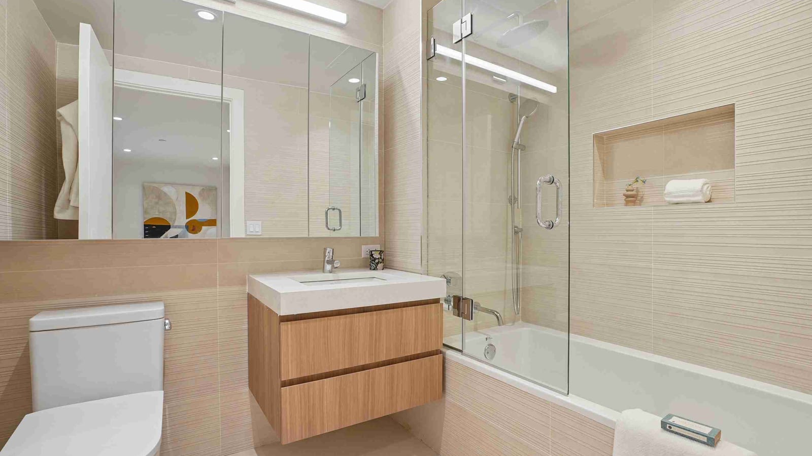 A bathroom with beige striped wall tile, a floating wood-grain vanity with a white sink, a wide mirrored cabinet, a toilet, and a bathtub with a glass shower screen and a wall niche holding a rolled towel