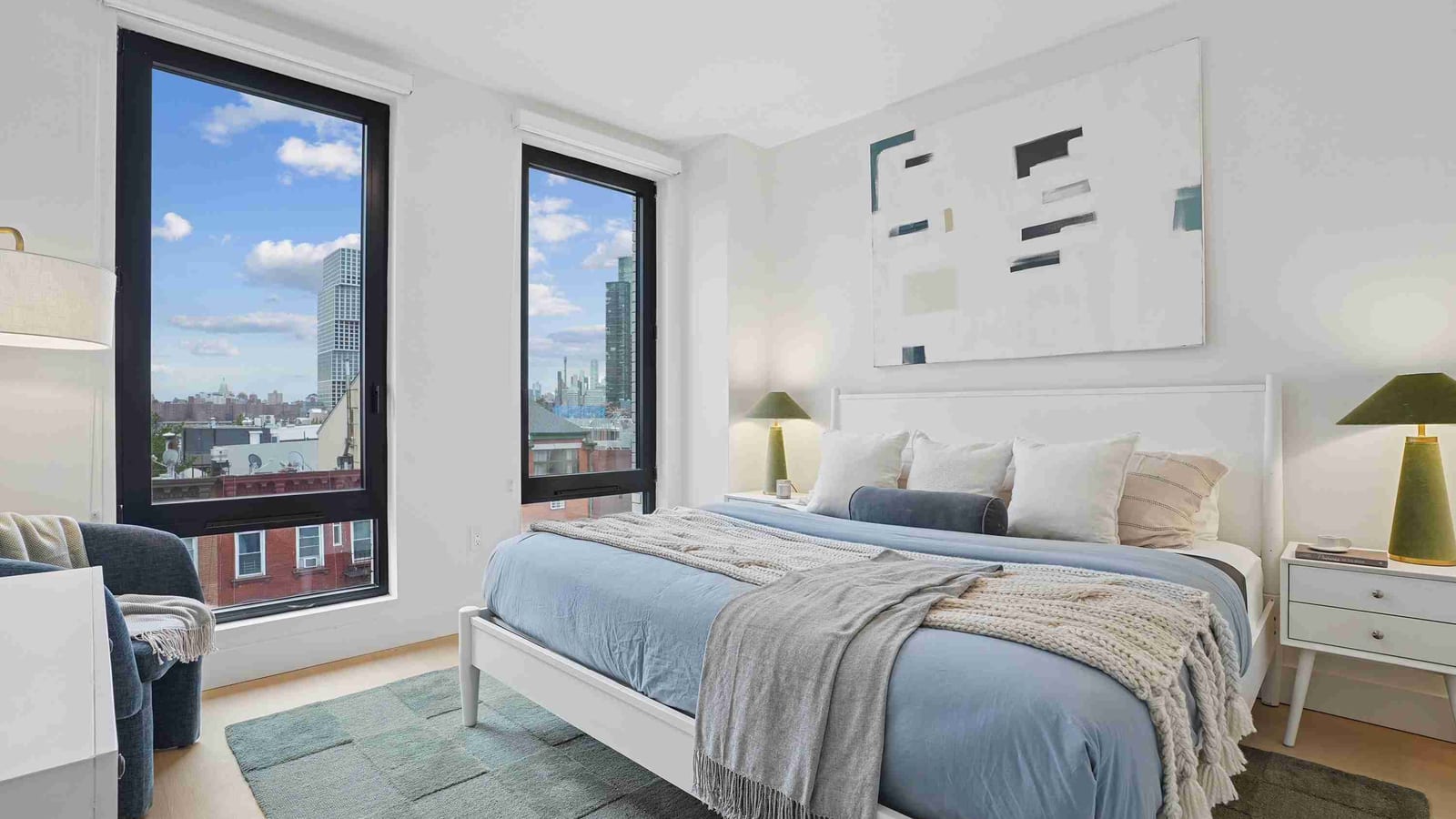 A bedroom with a white bed made up in blue and gray, two green bedside lamps, an abstract painting, a green rug, and two tall black-framed windows showing brick rowhouses and distant towers under a blue sky