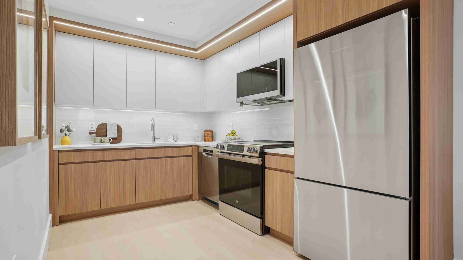 An L-shaped kitchen with white upper cabinets, wood-grain lower cabinets, a white stone counter with a sink, a stainless steel electric range with a microwave above, a dishwasher and a tall stainless steel refrigerator, lit by a strip of light along the ceiling