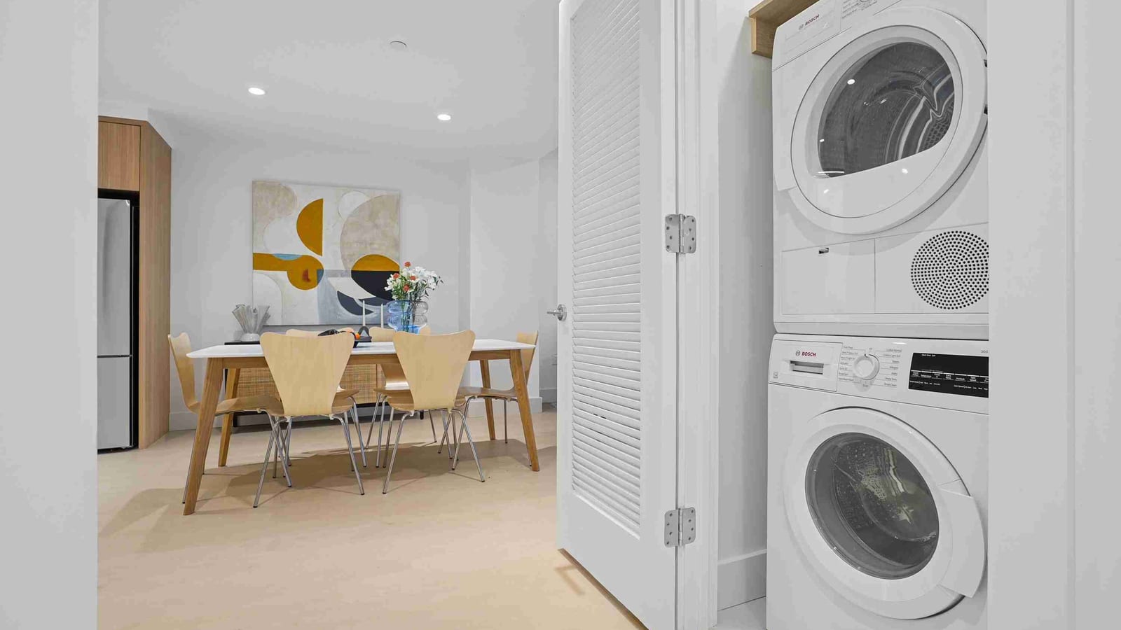 A white stacked Bosch washer and dryer in a closet with a louvered white door, beside a dining area with a wood table, four wood chairs, a vase of flowers and an abstract painting