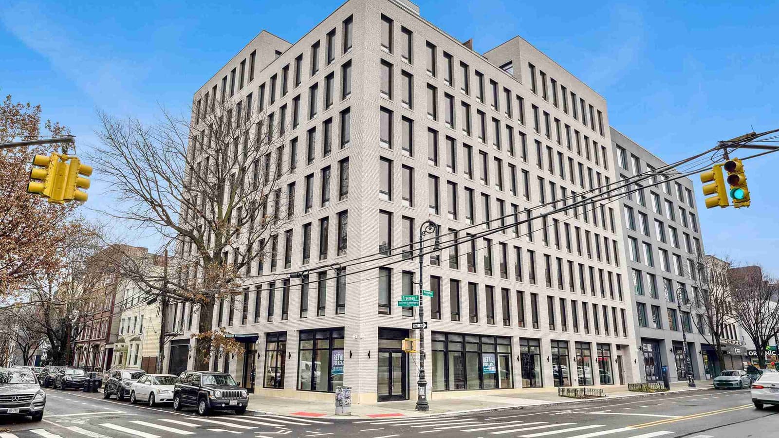 An eight-story light gray brick building on a street corner, with rows of tall dark-framed windows, glass storefronts with Retail For Lease signs at street level, green street signs reading Freeman and Manhattan on a lamppost, yellow traffic lights on overhead wires, bare trees and parked cars under a blue sky