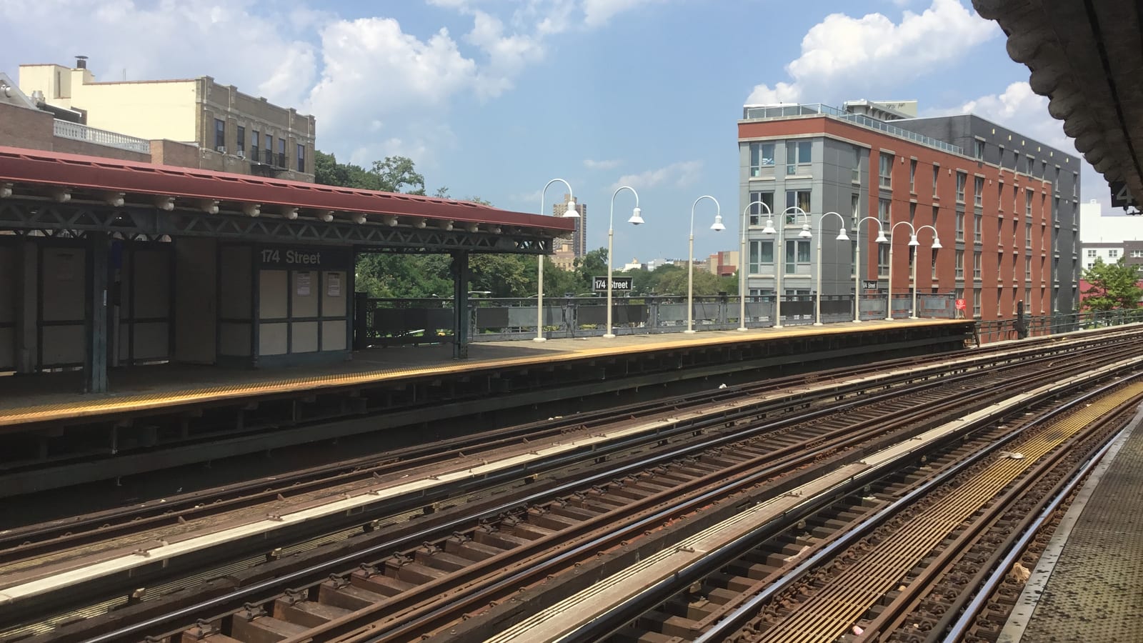Photograph from an elevated subway platform across several tracks to the opposite platform, with a red-roofed shelter and a sign reading 174 Street, white lamp posts, trees and apartment buildings behind, under a partly cloudy sky