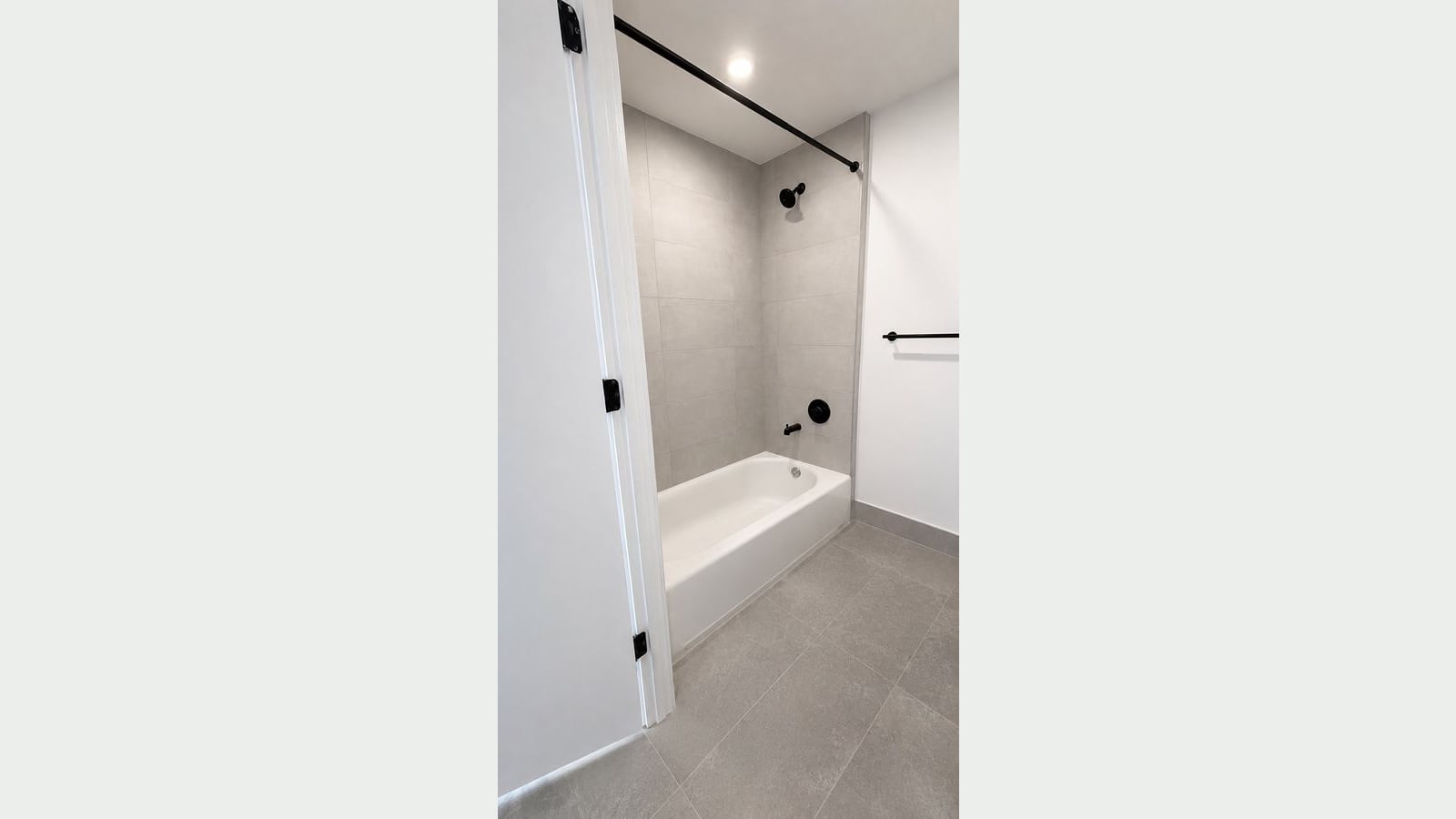 Photograph of a bathroom with a white tub, gray tile on the tub walls and floor, black shower fittings and a black curtain rod and towel bar, seen through an open white door