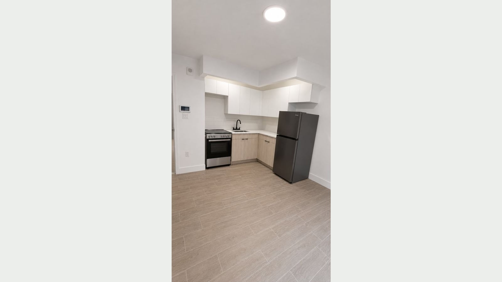 Photograph of an empty apartment with wood-look plank floors and an L-shaped kitchen: white upper cabinets, light wood lower cabinets, a white counter with a black faucet, a stainless electric range with a smooth cooktop, and a dark steel refrigerator, with an intercom panel on the wall at left