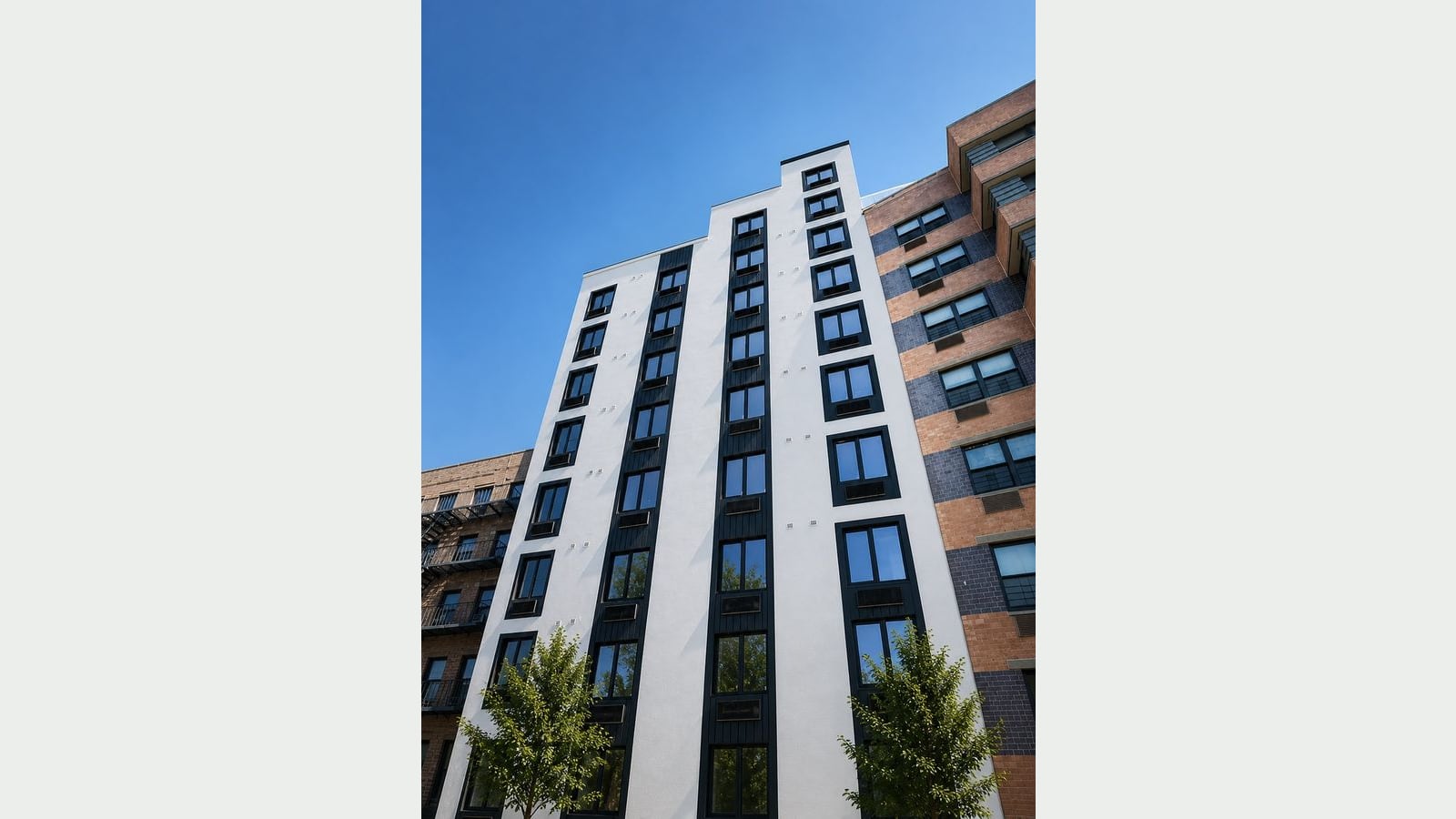 Photograph looking up at a nine-story building with a smooth white facade, black-framed windows with wall air-conditioner grilles, two center columns of windows set in black vertical panels, and a stepped top, between an older brick building with fire escapes and a newer orange and dark blue striped brick building, with two young street trees and a blue sky