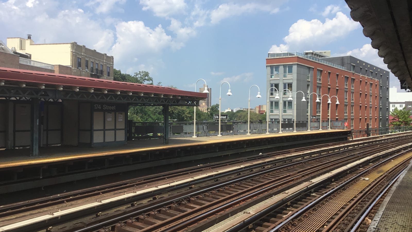 Photograph from a raised subway platform looking across four sets of tracks to a second platform with a red canopy and a black sign reading 174 Street, a row of white lamp posts, trees, and a gray and red brick apartment building under a blue sky with white clouds