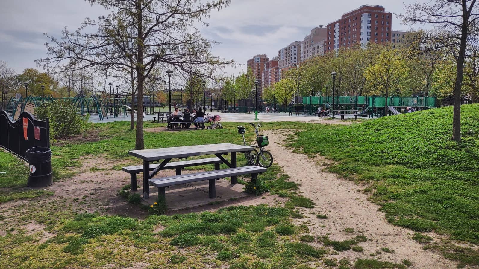 Photograph of a park lawn with a gray picnic table and a parked folding bicycle in front, a dirt path, green play equipment and a few small distant figures at tables behind, and tall brick apartment towers beyond the trees under a cloudy sky