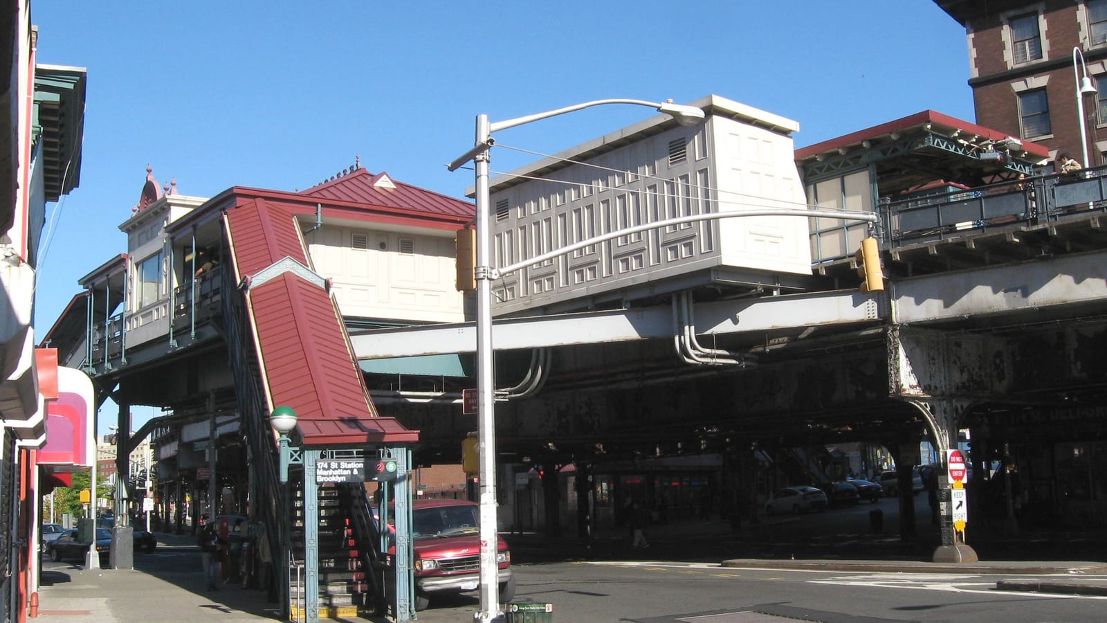 Photograph of a raised subway station over a wide street, with a red-roofed stair down to the sidewalk under a sign reading 174 St Station, Manhattan and Brooklyn, 2 and 5, a gray station house on steel girders, a red pickup truck at the curb and a brick apartment building at the right, under a clear blue sky