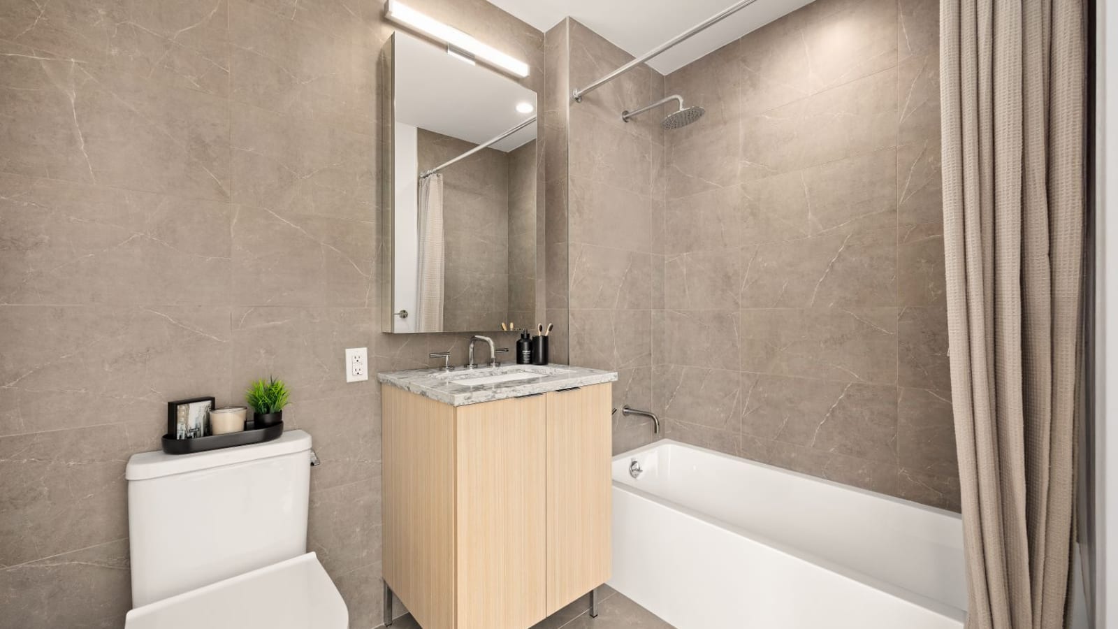Photograph of a bathroom with large gray stone-look tiles, a light wood vanity with a marble-look top, a mirror cabinet with a light bar, a white toilet and a tub with a rain shower head and a beige curtain