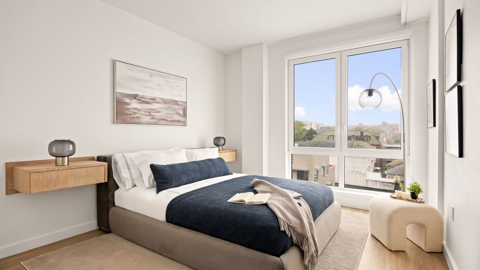 Photograph of a furnished bedroom with a gray upholstered bed and navy blanket, floating wood nightstands with lamps, an arc floor lamp, and a large window looking over low rooftops and trees