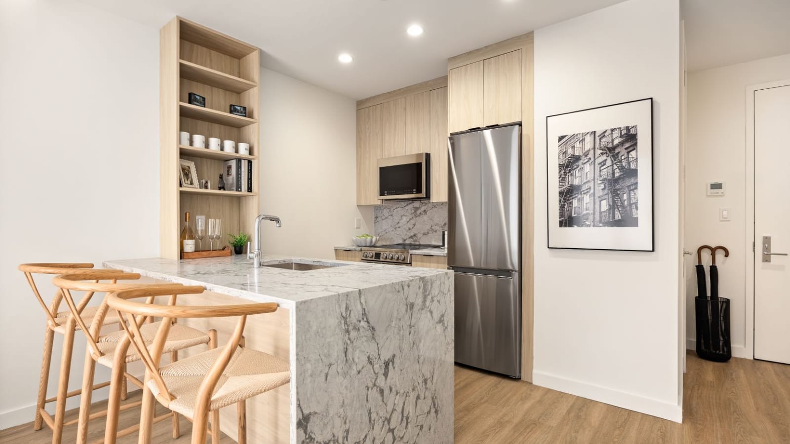 Photograph of an open kitchen with light wood cabinets, a gray and white marble-look counter with three wood stools, a stainless range with a flat top and a microwave above, a stainless refrigerator and an open shelf unit