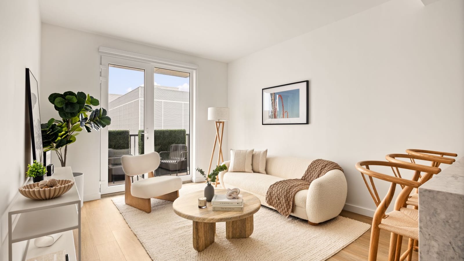Photograph of a furnished living room with a cream sofa, a round wood coffee table, a floor lamp and wood-look floors, and glass doors opening to a small outdoor space with hedges and a chair