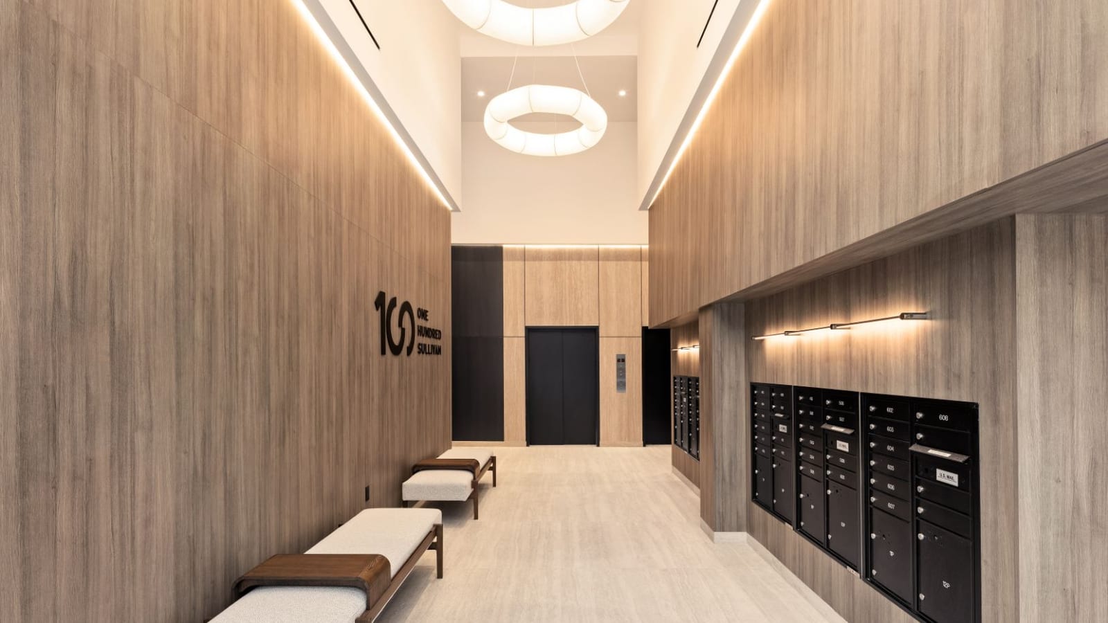 Photograph of a tall lobby with wood-look walls, a sign reading 100 One Hundred Sullivan, two upholstered benches, ring-shaped ceiling lights, black mailboxes along the right wall and a black elevator door at the end