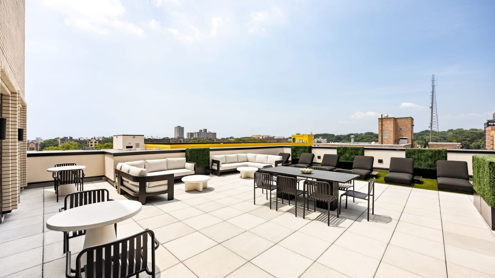 Photograph of a paved roof terrace with outdoor sofas, a long dining table with chairs, lounge chairs, boxed hedges and a strip of artificial grass, with low buildings, trees, a radio tower and a few apartment towers in the distance