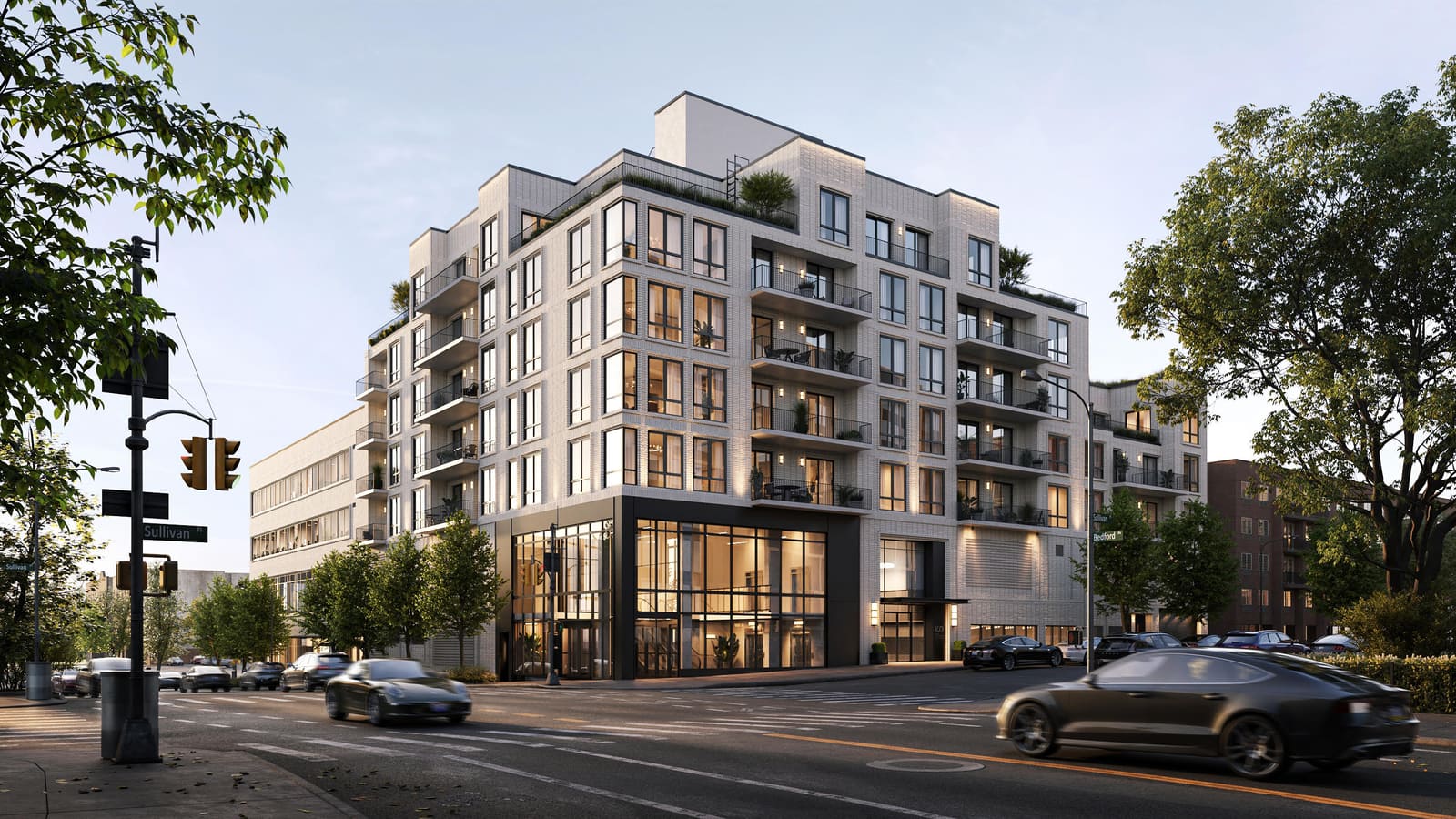 Rendering at dusk of a pale brick apartment building on a street corner, with balconies, a double-height glass storefront at the corner, a lower office-style wing at left, street signs for Sullivan Place and Bedford Avenue, and two dark cars driving past