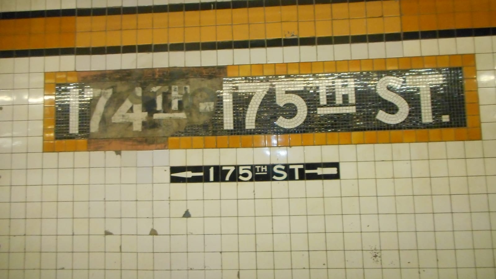 Photograph of a white tiled subway wall with an orange and black border and a mosaic name panel reading 174th-175th St., the 174th part worn and patched, above a small black mosaic sign reading 175th St with arrows