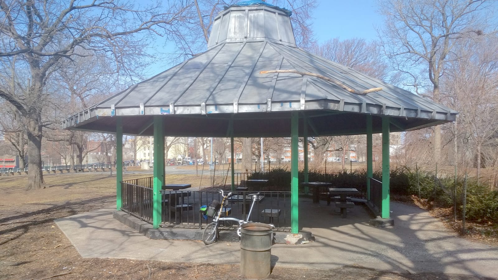 Photograph of an octagonal park pavilion with a grey metal roof and green posts over stone game tables, a folding bicycle and a metal trash can in front, bare trees and apartment buildings behind under a blue sky