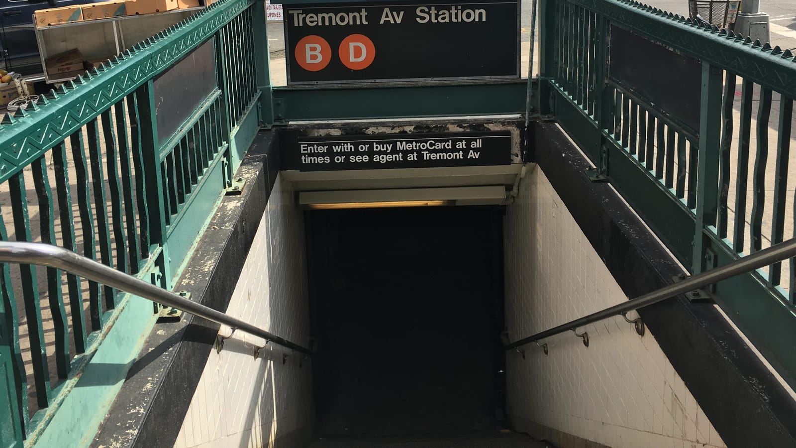 Photograph of a subway stair entrance with green railings, a black sign reading Tremont Av Station with orange B and D route circles, and a second sign about MetroCards over steps going down