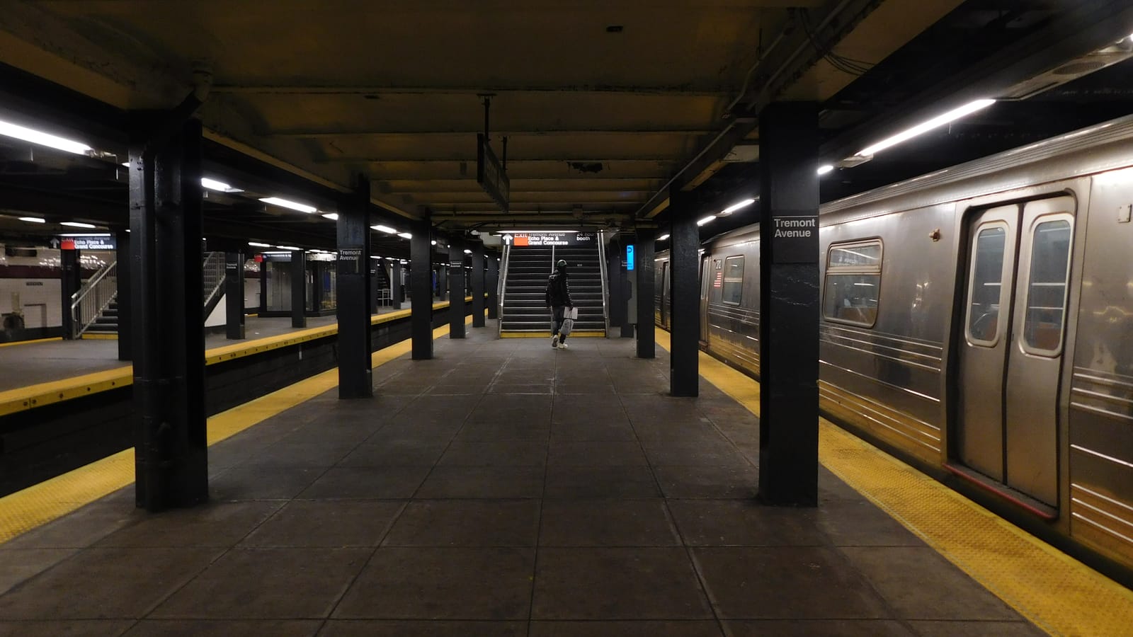 Photograph of an underground subway platform with black columns marked Tremont Avenue, yellow platform edges, a stair at the far end with an exit sign, one person walking away, and a silver train with closed doors on the right