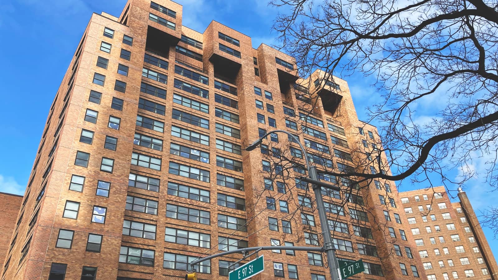 Photograph looking up at a tall orange-brown brick apartment tower with bands of dark windows and four boxy brick blocks projecting near the top, under a blue sky with thin clouds, with bare tree branches at the right, a street light on a pole and green street signs reading E 97 St at the bottom edge