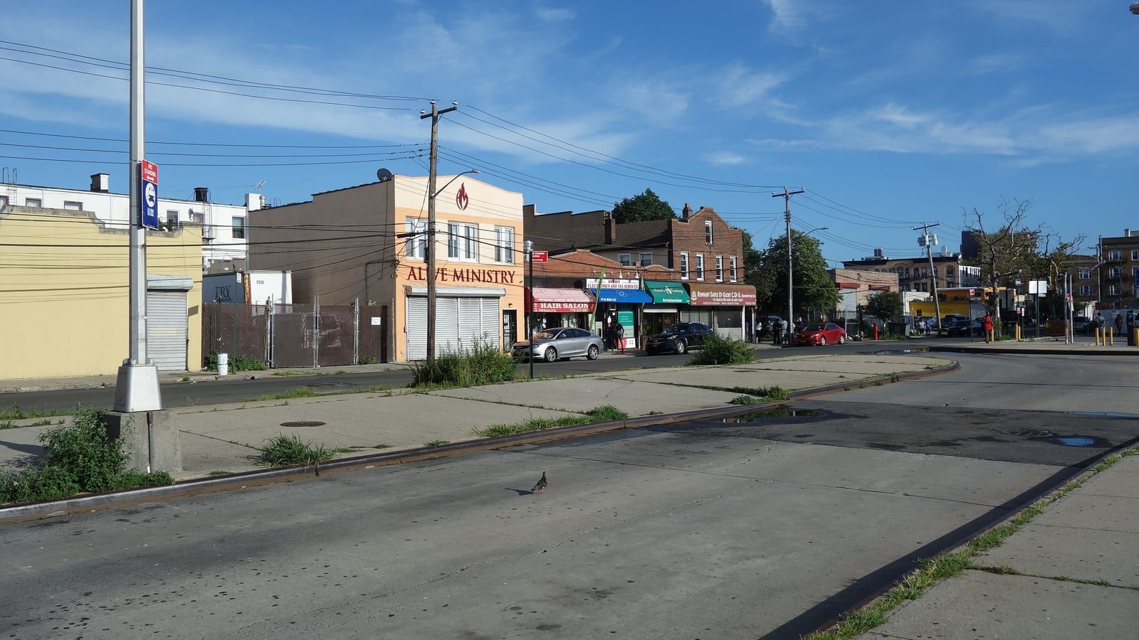 Photograph of a wide, empty bus lane beside a sidewalk with a tall light pole and a bus stop sign, and across the street a row of low buildings with shop fronts, a yellow building with a closed shutter and a beige building with a sign reading Alive Ministry, under a blue sky with thin clouds