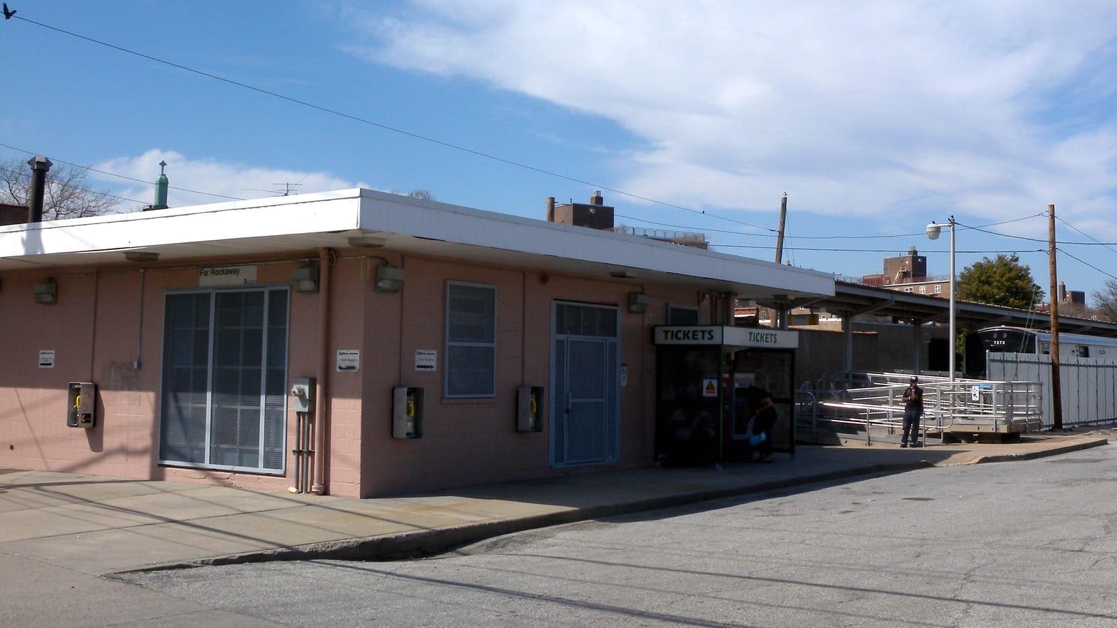 Photograph of a one-story pink station building with a flat white roof, a small sign reading Far Rockaway, pay phones on the wall, a green Tickets booth, a ramp with metal railings leading to a covered platform and the end of a train at right, and apartment buildings behind under a blue sky with clouds