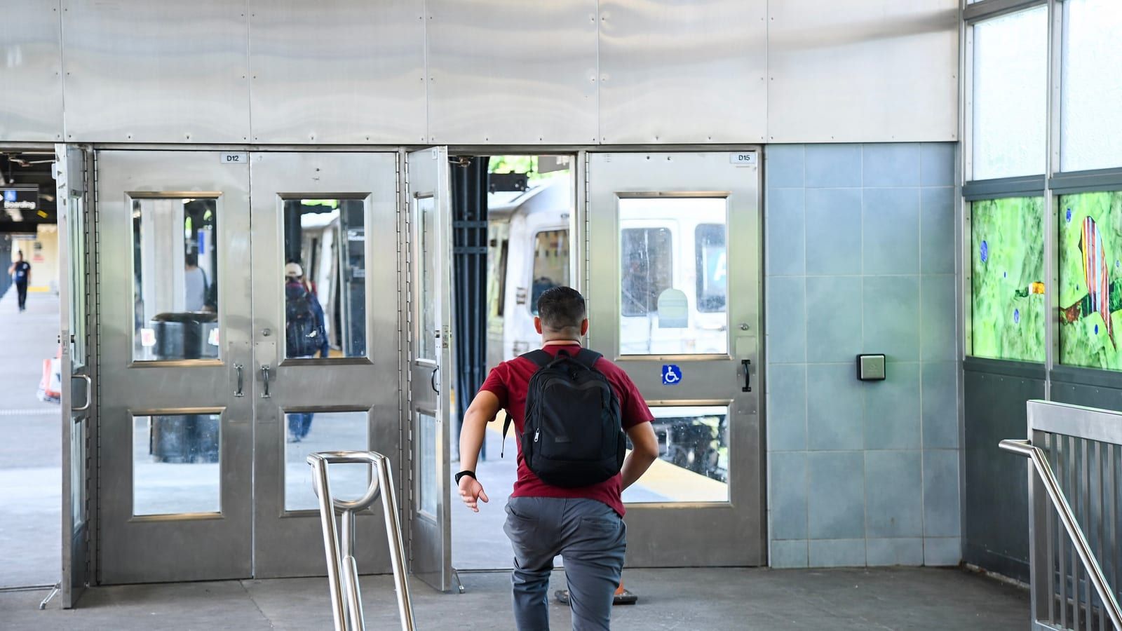 Photograph inside a subway station of brushed steel walls and two sets of steel doors with glass panes, one door open onto a platform where a silver train waits, a man in a red shirt with a black backpack walking toward it seen from behind, a steel handrail in front and colorful glass artwork panels at right