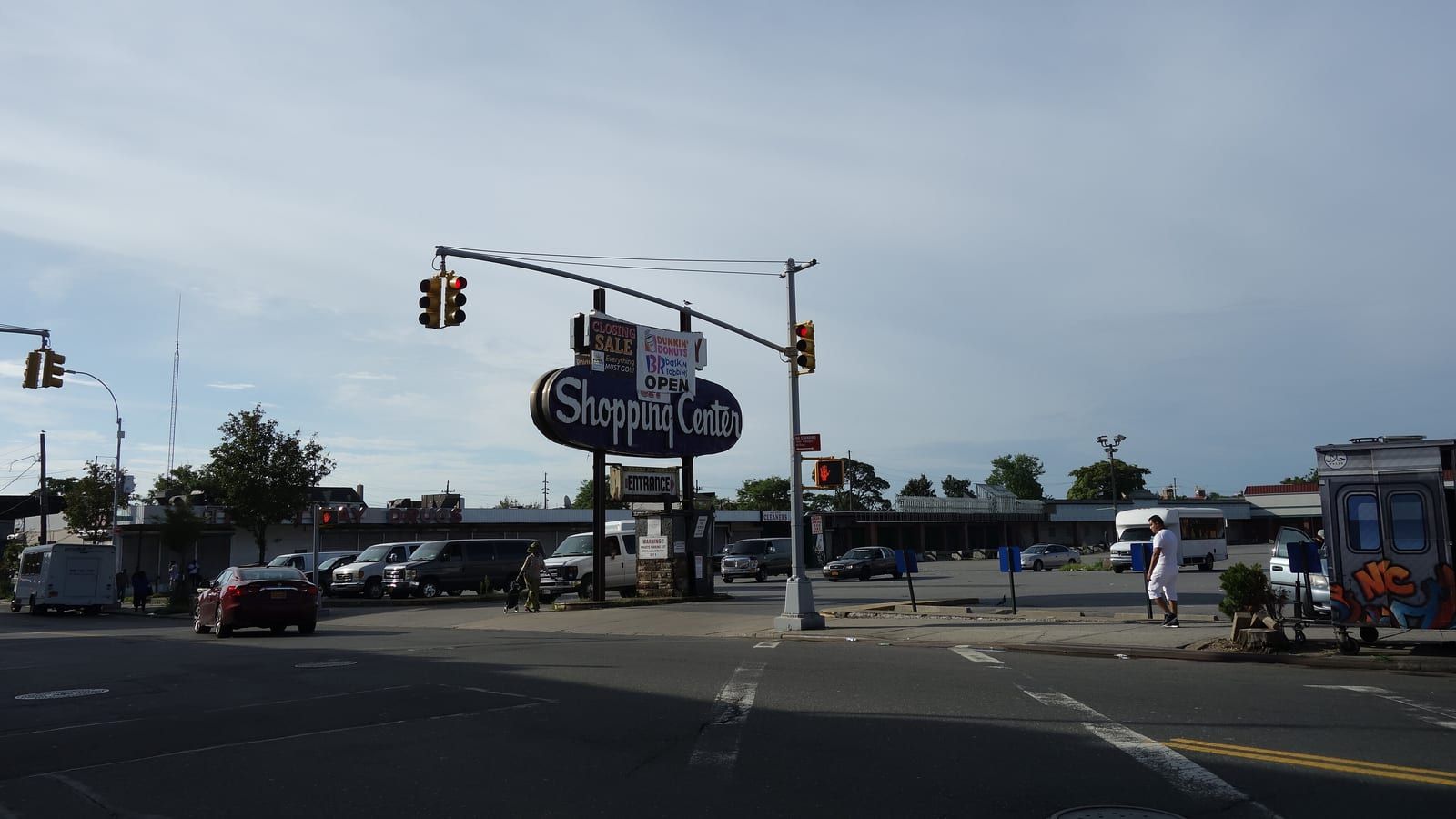 Photograph of a wide street intersection with traffic lights on a curved pole, a tall oval sign reading Shopping Center over a parking lot full of cars and a row of low stores behind it, a few people walking at a distance, under a pale sky