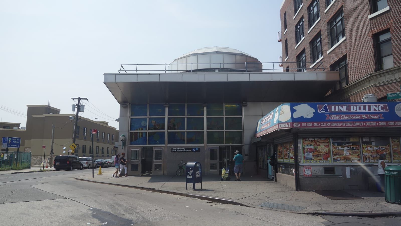 Photograph of a low subway station house with a flat overhanging roof, a round skylight dome on top, tall windows of colored glass over two sets of doors and a sign reading Far Rockaway Mott Avenue Station with the A train symbol, a blue mailbox on the sidewalk, a deli with a blue awning at right, a brick apartment building behind it and a beige building and parked cars down the street at left, under a hazy sky