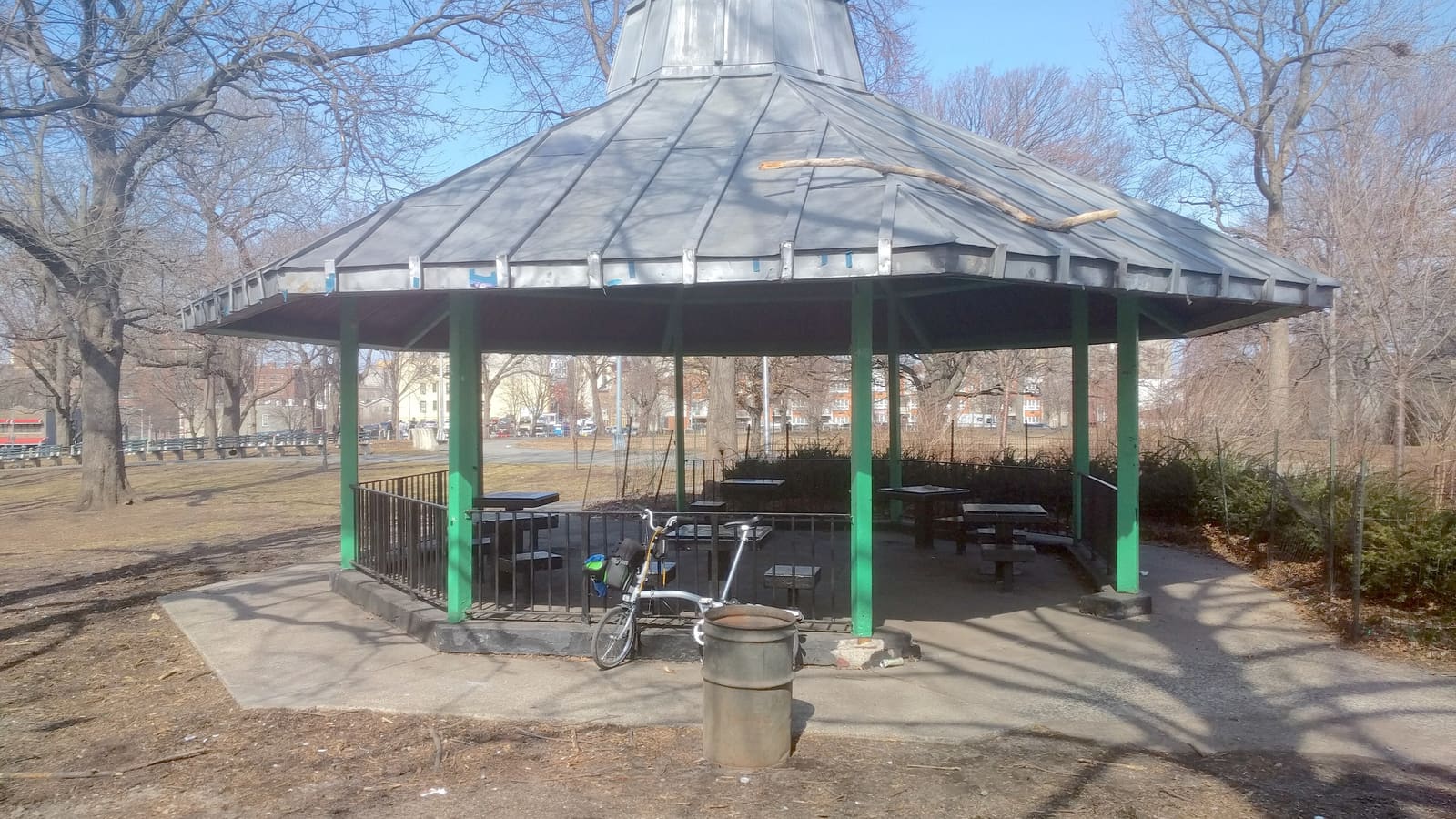 An open-sided gazebo with a gray metal roof on green posts, with game tables inside a black railing, a folding bicycle parked against it, bare trees and apartment buildings in the background