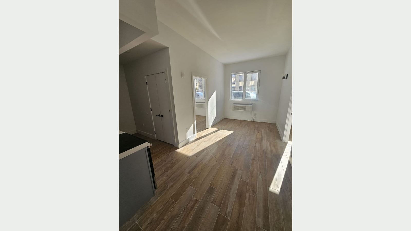 Photograph of an empty, sunlit room with wood-look plank flooring, white walls, a wall heating and cooling unit under a window, a double closet door at left, an open door to a second room and the edge of a kitchen counter with a black electric cooktop at lower left