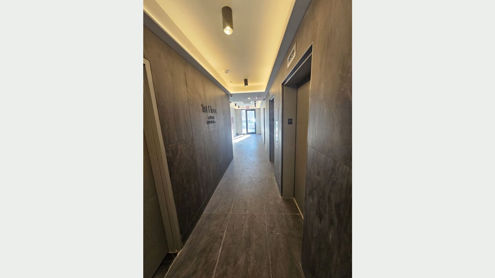 Photograph of a ground-floor hallway with dark stone-look wall and floor tiles, lit ceiling cove and spotlights, a sign reading 1st Floor with apartment arrows, two elevator doors at right and a glass entrance door at the far end