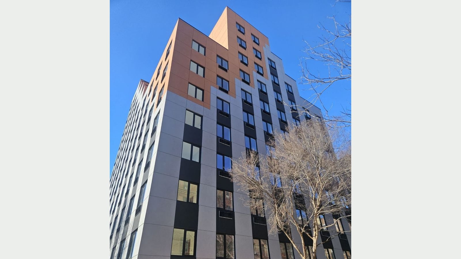 Photograph looking up at a 12-story apartment building under a clear blue sky: light gray panels with black bands around rows of windows, an orange-brown upper section that steps up at the top, wall vents under many windows and a bare tree in front at right