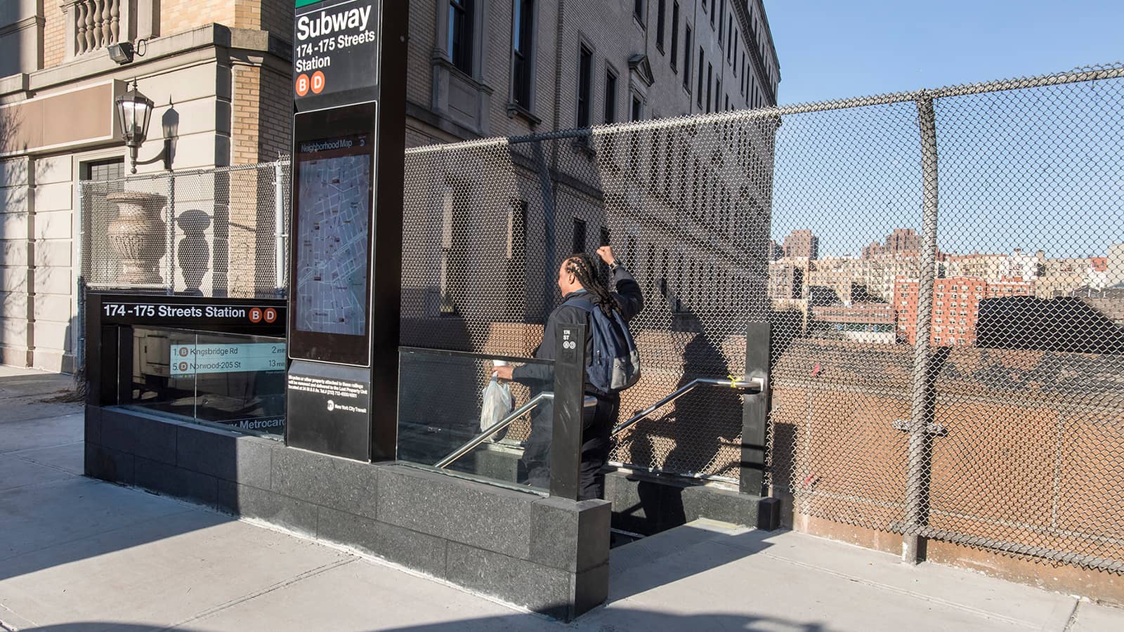 Photograph of a new black granite subway entrance with a tall sign reading Subway 174-175 Streets Station with B and D symbols and a neighborhood map, beside a chain-link fence above an open cut, with a person seen from behind going down the stairs and apartment buildings in the distance