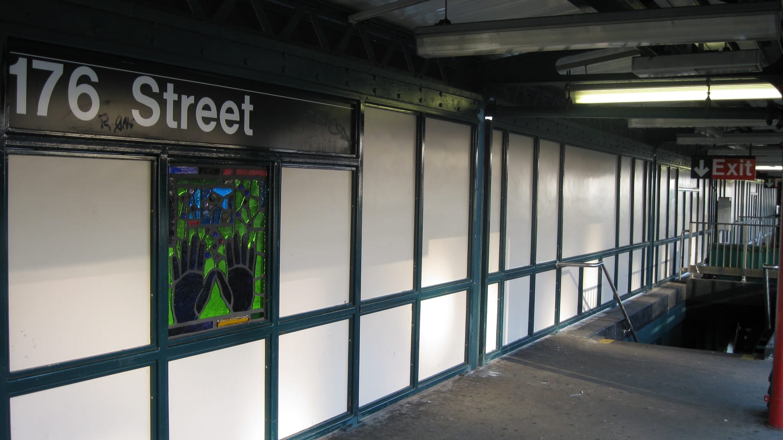 Photograph of an elevated subway platform with a black sign reading 176 Street above white wall panels in green frames, a stained-glass panel showing two hands, fluorescent lights and a red Exit sign above the stairs