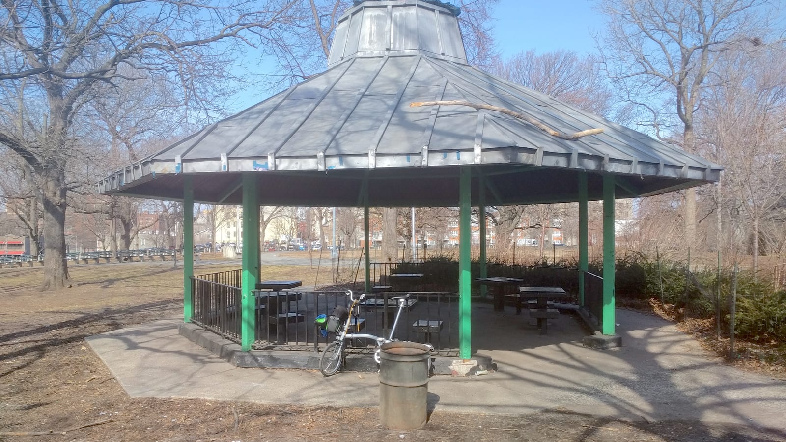 Photograph of an eight-sided park shelter with a gray metal roof on green posts, with stone tables and benches inside, a parked bicycle and a trash can in front, bare trees and apartment buildings in the background
