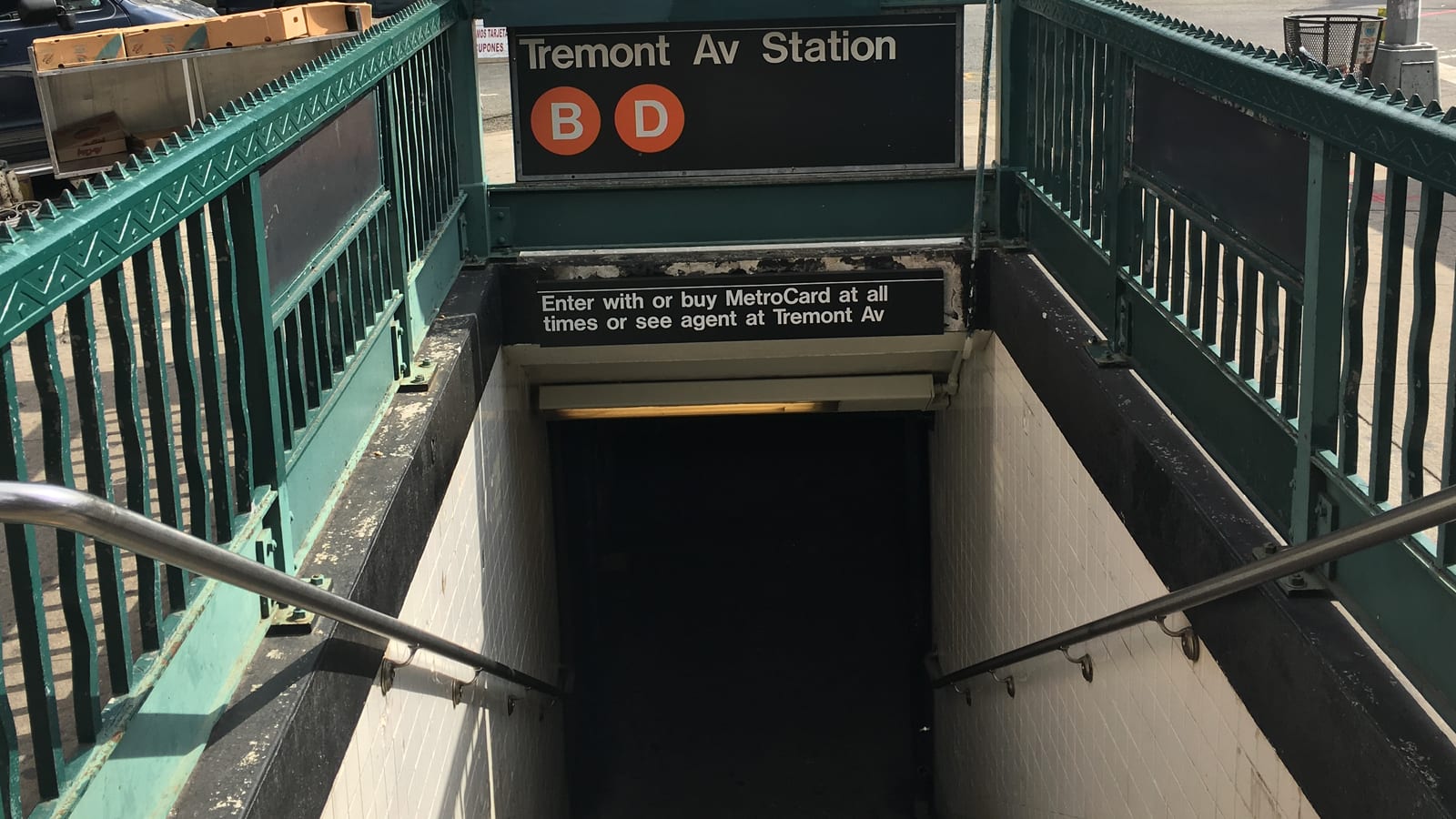 Photograph looking down a subway entrance stair between green iron railings, under a black sign reading Tremont Av Station with orange B and D symbols and a smaller sign about MetroCards, with a sidewalk and parked van beyond