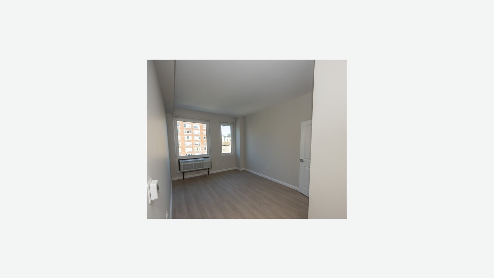 An empty bedroom with gray walls, wood-look flooring, a white door, a thermostat on the left wall and two windows, one with a wall air conditioner below it, looking out at a brick apartment building