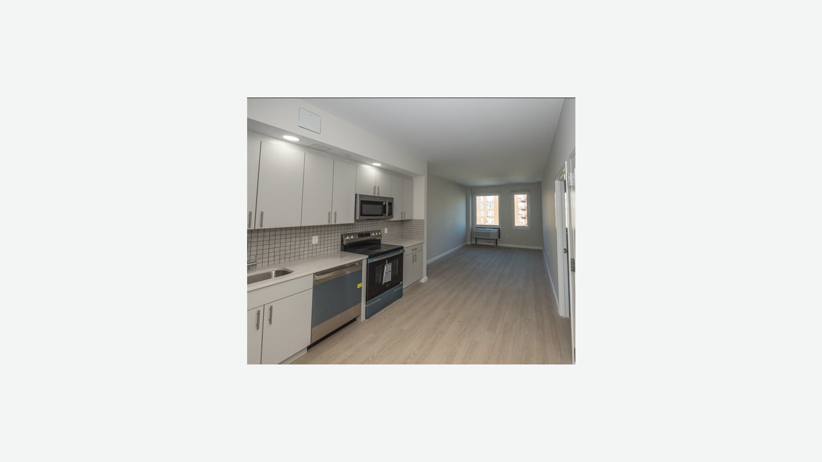 A long open room with white upper and lower cabinets, a gray square-tile backsplash, a stainless steel microwave over a black electric range with a smooth top, a dishwasher with a blue front, and light wood-look flooring leading to two windows with a wall air conditioner beneath