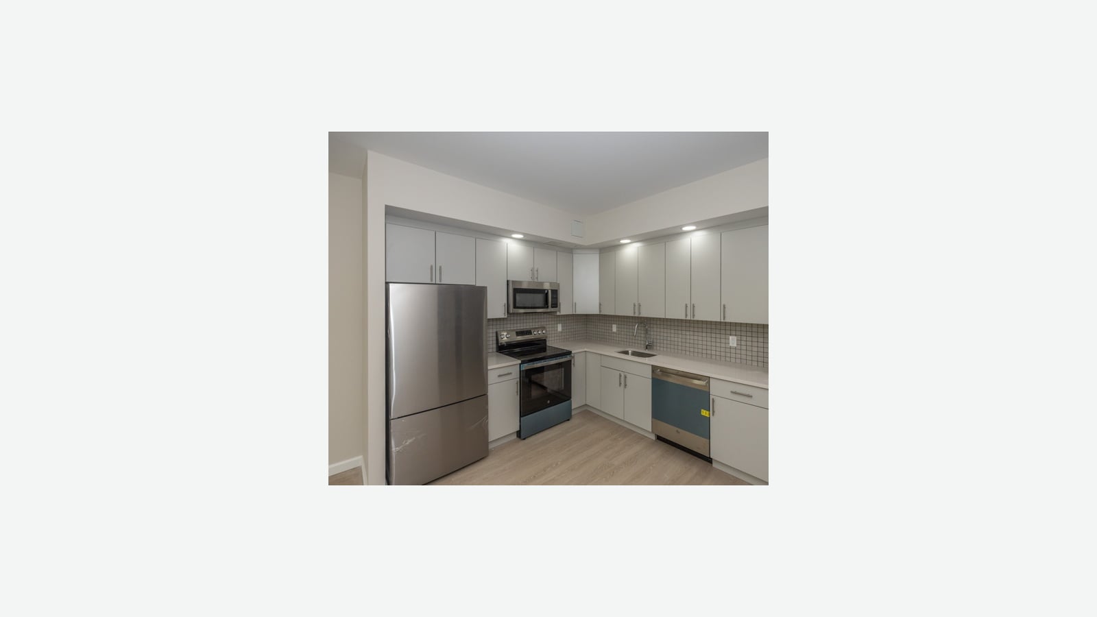 An L-shaped kitchen with white flat cabinets, a stainless steel refrigerator still wrapped in plastic film, a microwave over a black electric range, a square-tile backsplash, a sink under the corner cabinets and a dishwasher