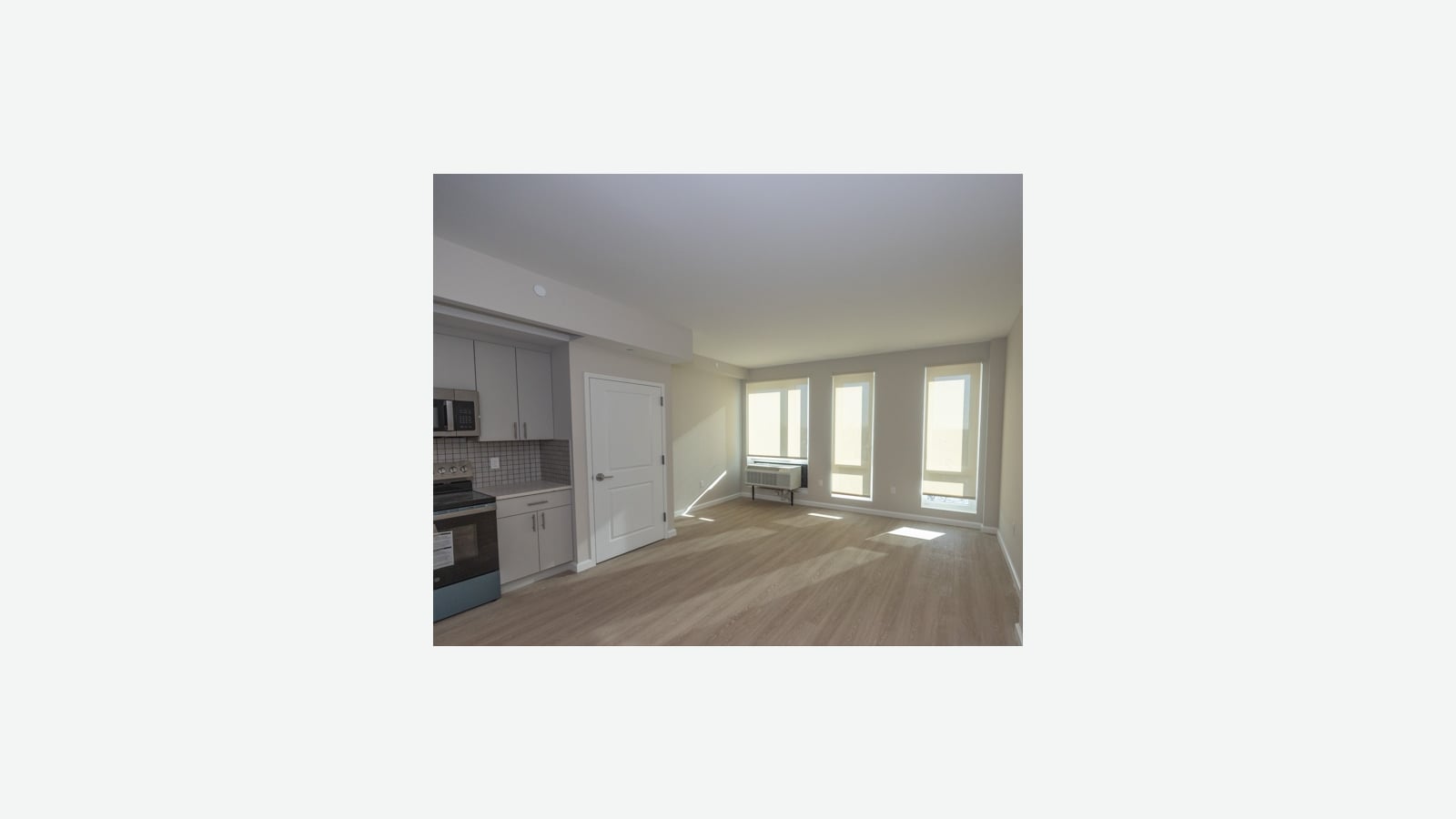 An empty room with light wood-look flooring and sunlight coming through three tall windows, a wall air conditioner under the left window, a white door and a kitchen corner with a microwave and a black range at left